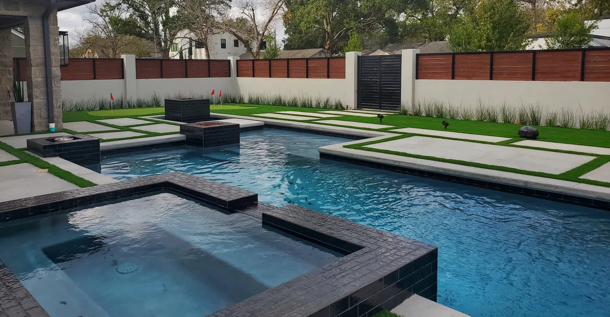 A dark modern pool and raised spa with turf-and-paver strips along the deck, inside a rendered wall