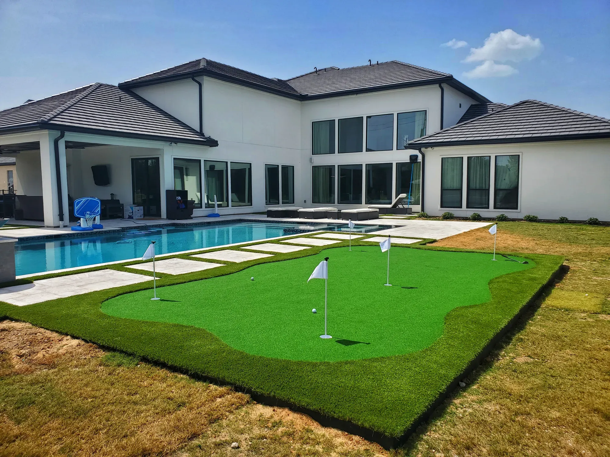 A rectangular backyard putting green with flags, laid beside a swimming pool and a modern home