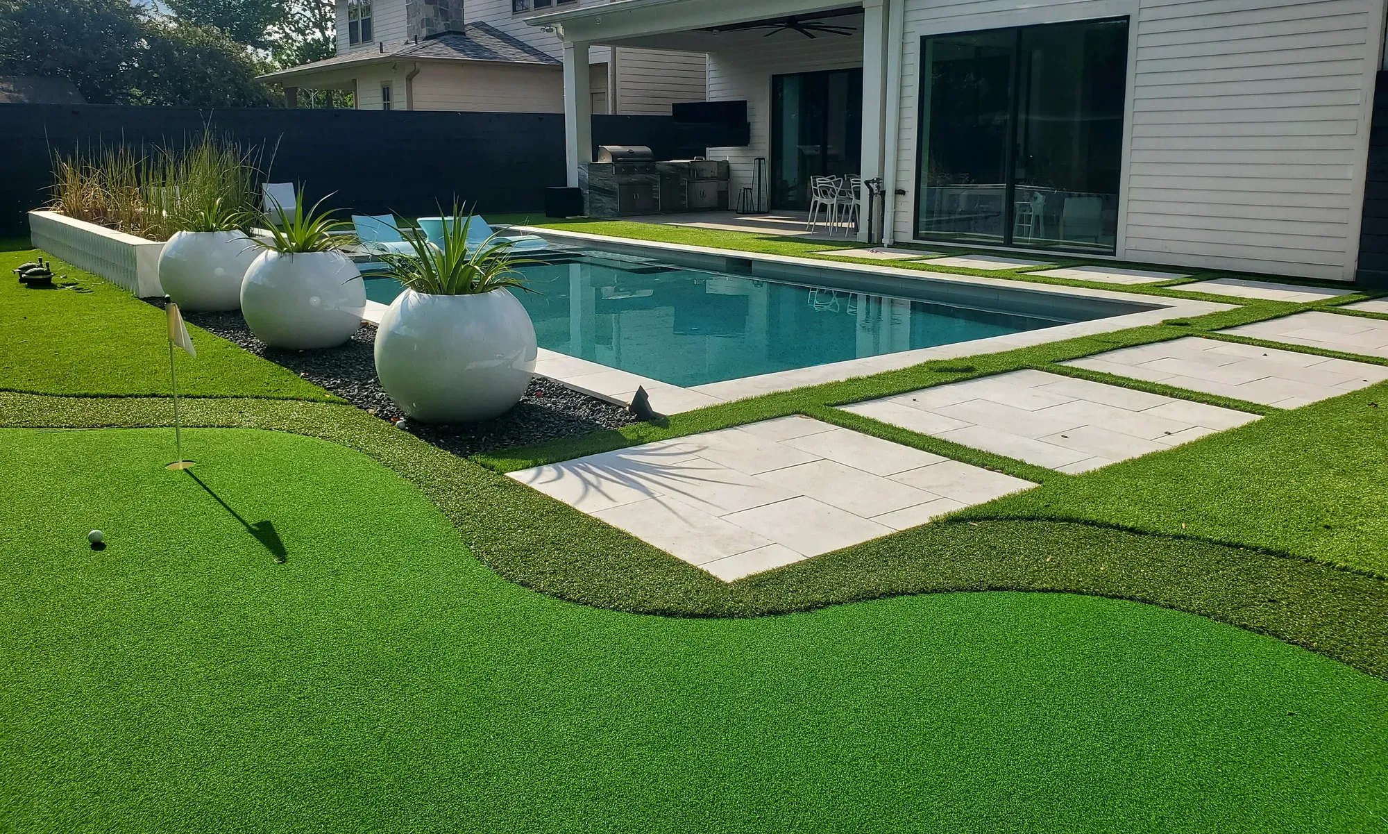 A pool deck of paver strips opening onto a broad curved artificial lawn with planters