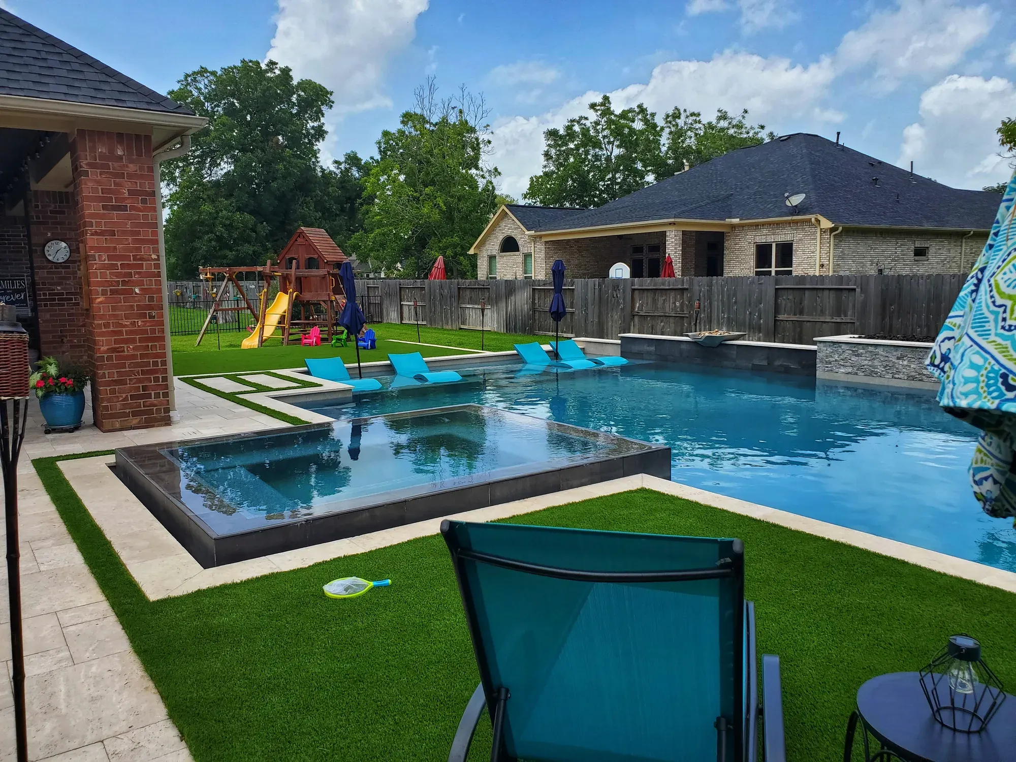A pool and raised spa surrounded by artificial turf, with a play structure and homes beyond