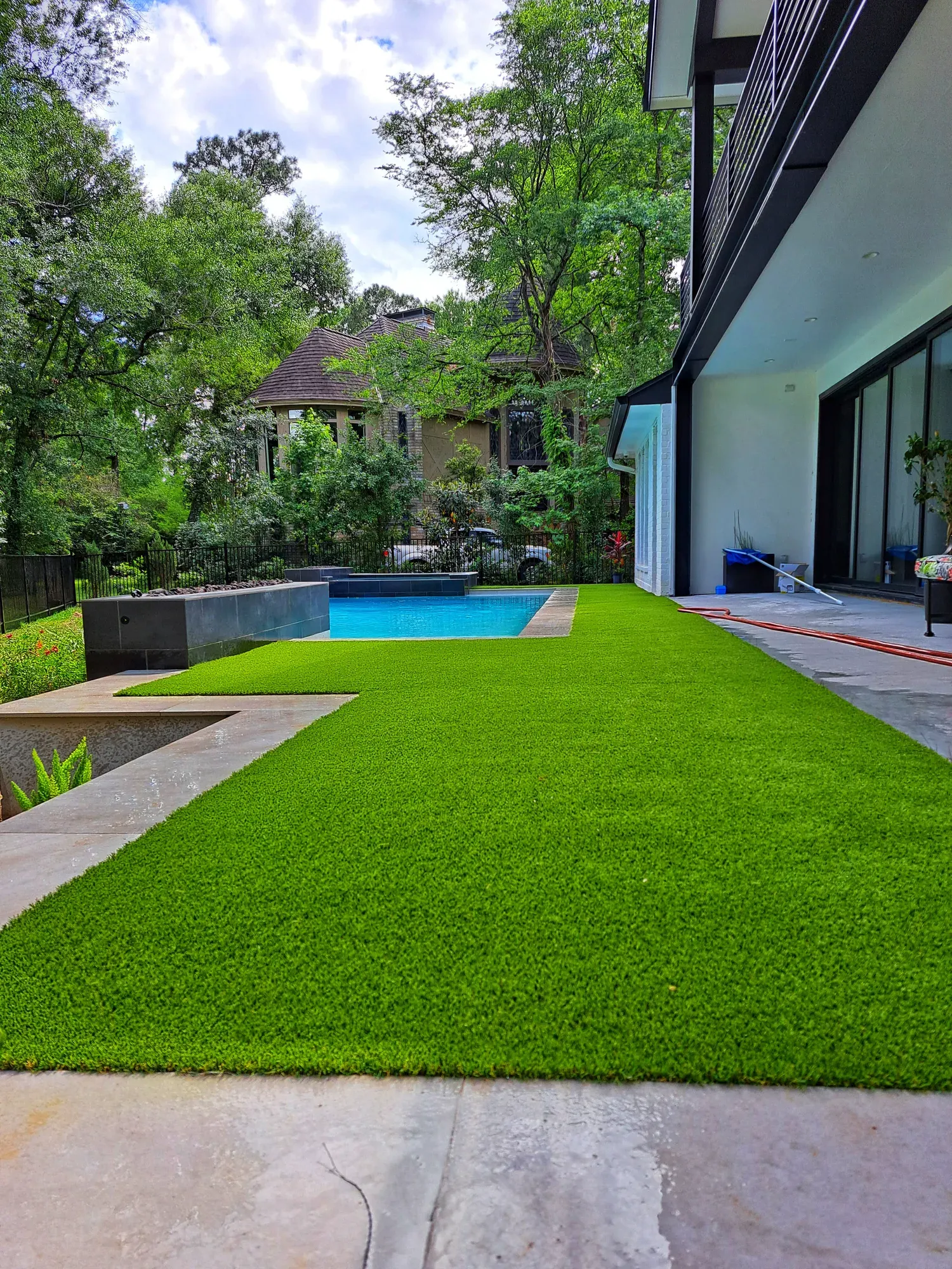 An artificial lawn running between a house wall and a raised pool in dense tree cover