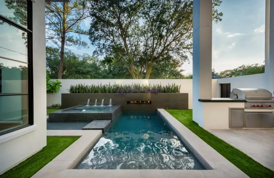 A narrow lap pool and spa with artificial turf along one side and an outdoor kitchen beyond