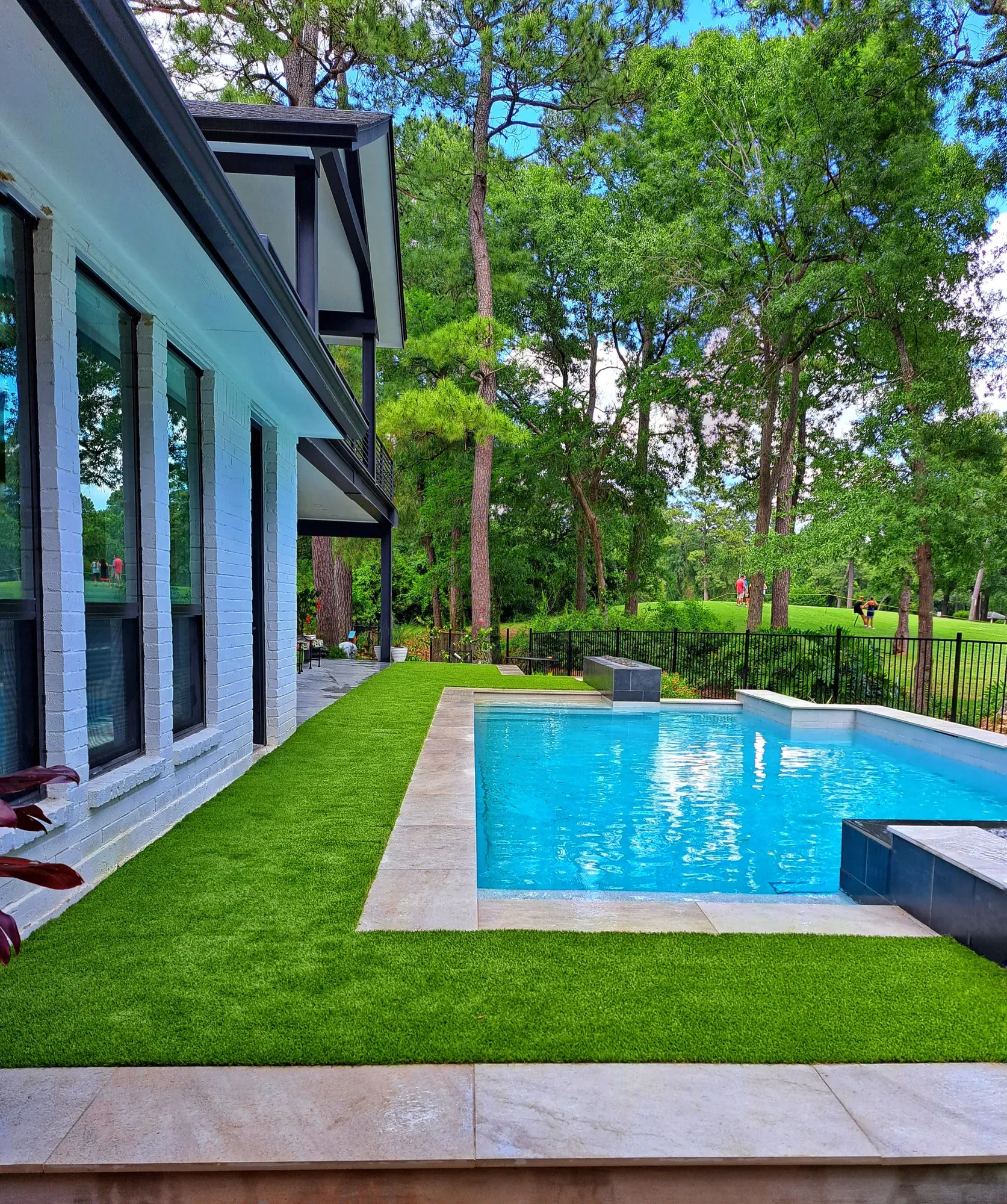 Strip of artificial turf running between a white brick house and a rectangular pool, with pine woods behind