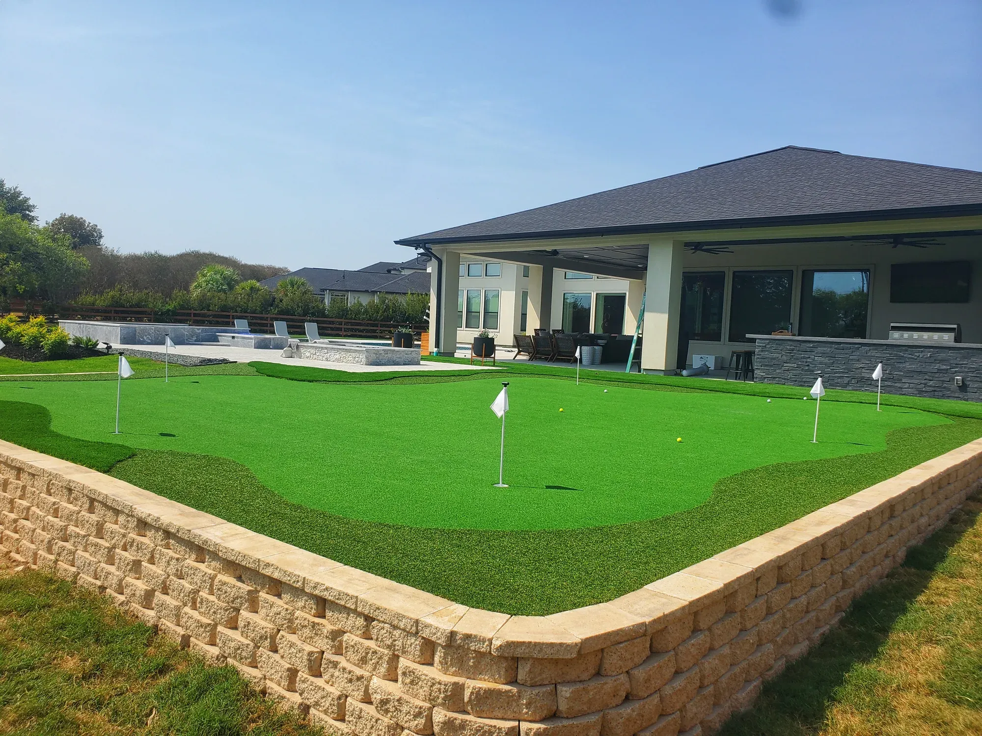 Artificial putting green with four flags built on top of a tan stone retaining wall behind a house