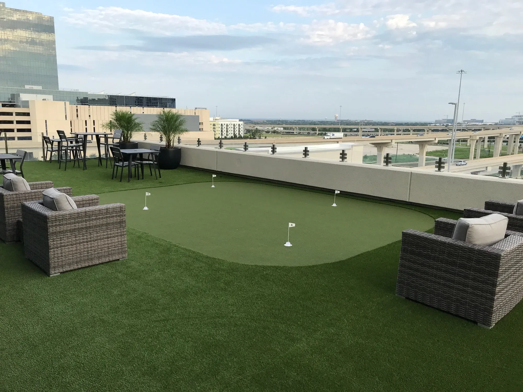 Artificial putting green with four cups and flags on a rooftop amenity deck, wicker lounge chairs around it and a freeway beyond the parapet