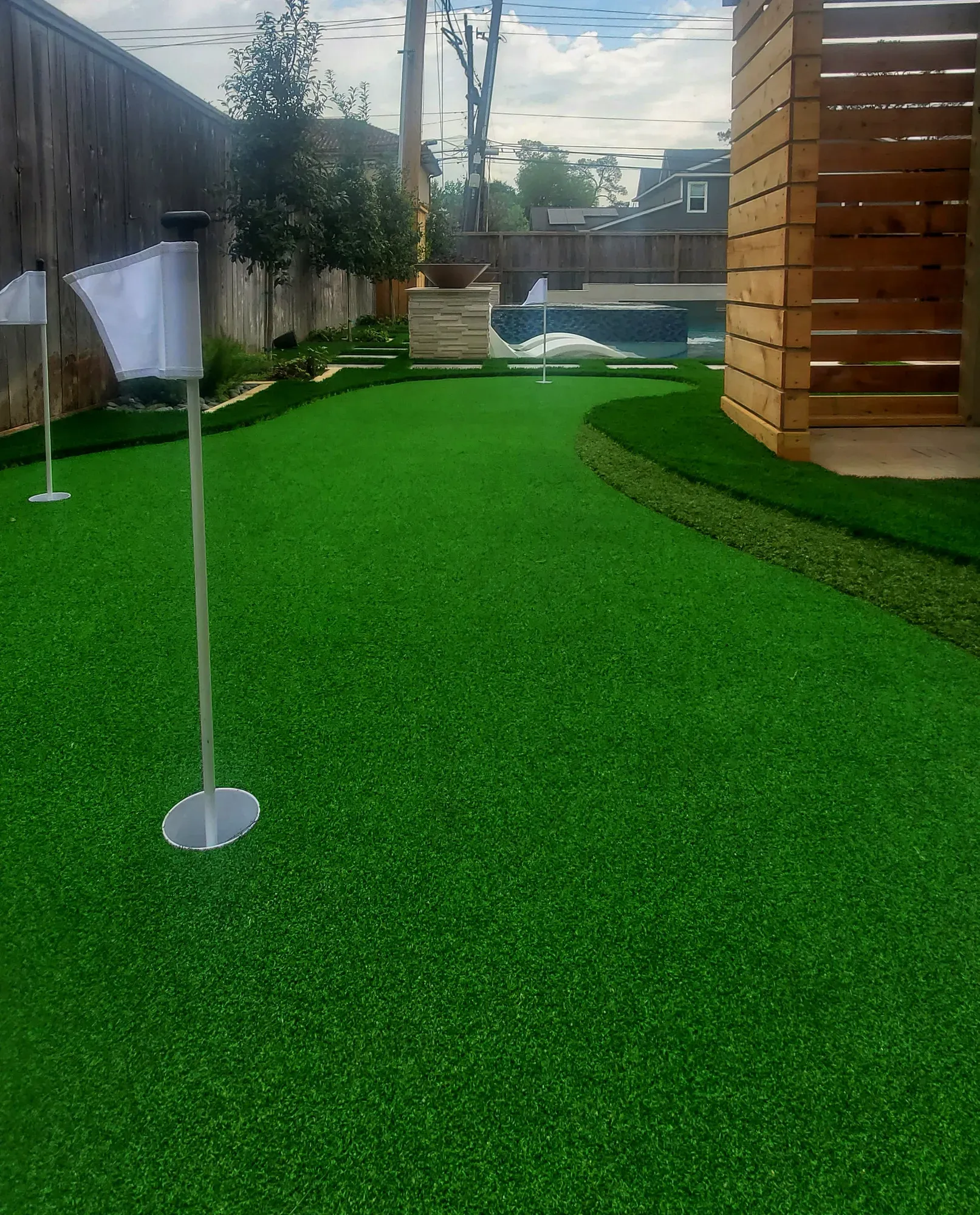 A side-yard practice green with a flag and cut-in cup, screened by a timber slat wall