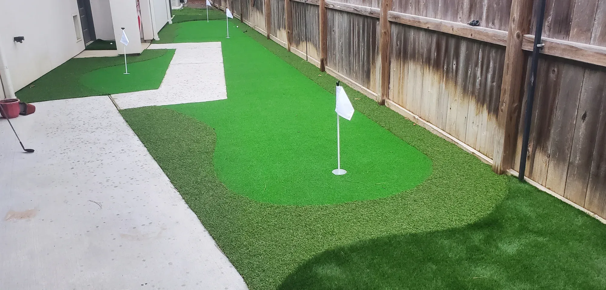A small side-yard practice green with a flag, set beside a concrete path and timber fence