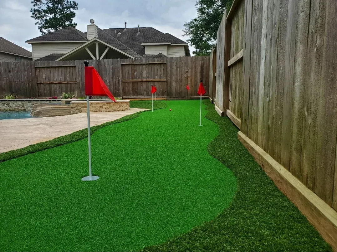 A narrow side-yard putting green with red flags against a timber fence
