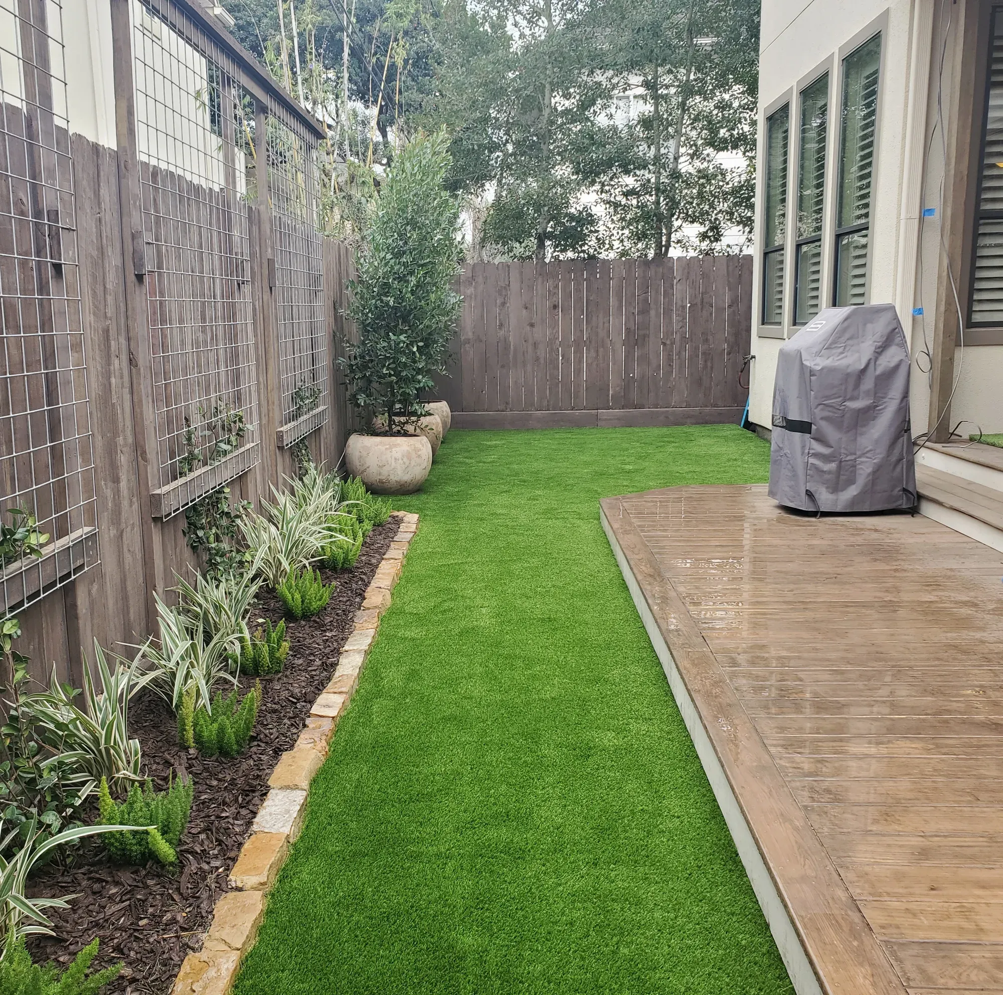 A narrow backyard laid in artificial turf beside a stained concrete deck and a planted border