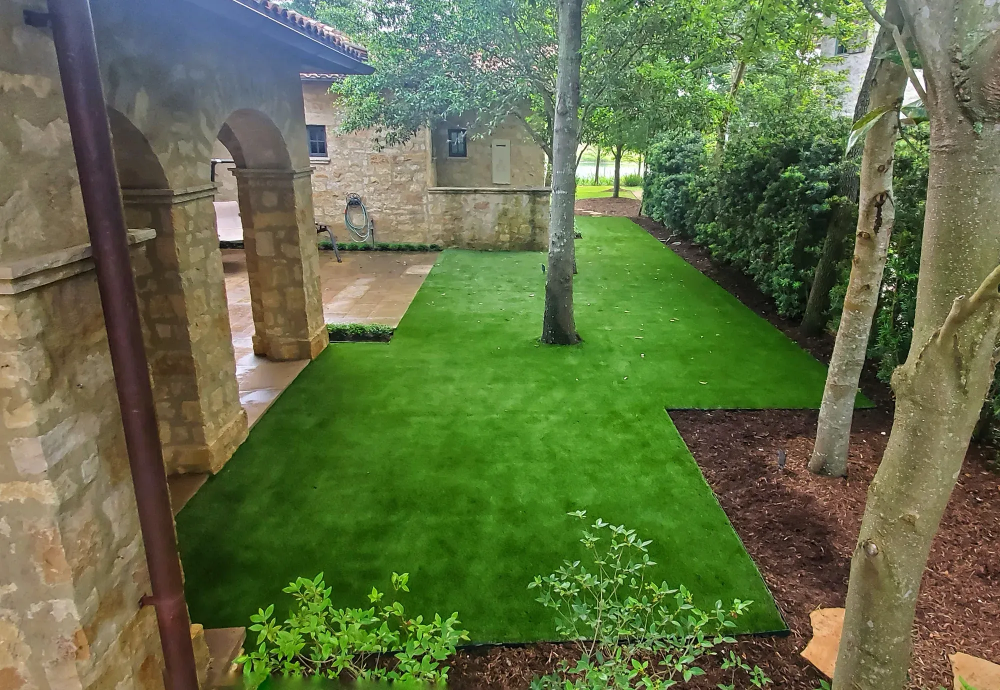 A shaded courtyard lawn of artificial turf running between a stone house and mature trees