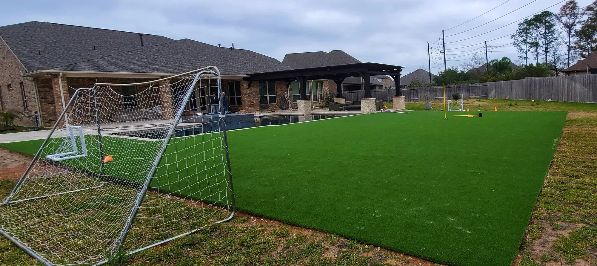 Artificial turf field with a full-size soccer goal standing on it, running back past training cones to a covered patio and pool behind a house