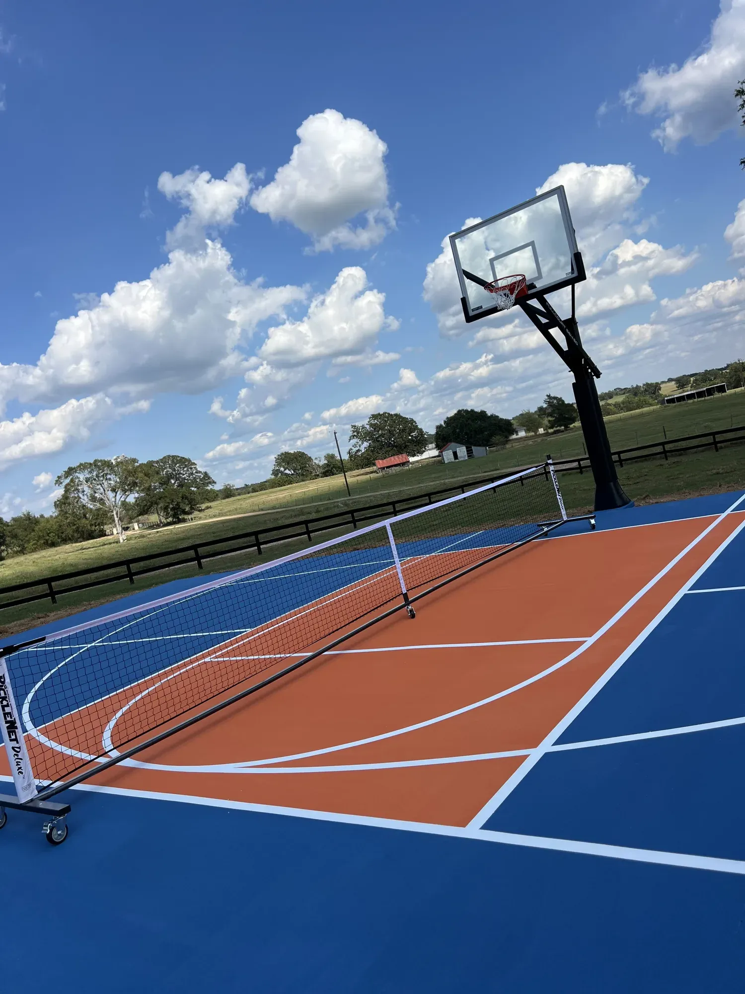 A portable pickleball net standing across an outdoor acrylic sport court, a basketball goal at the far end and open pasture beyond the fence