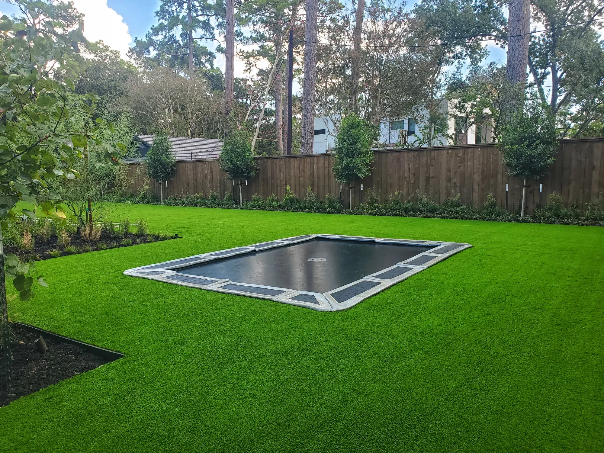 In-ground trampoline set flush into an artificial turf back yard, edged with stone and surrounded by fresh planting