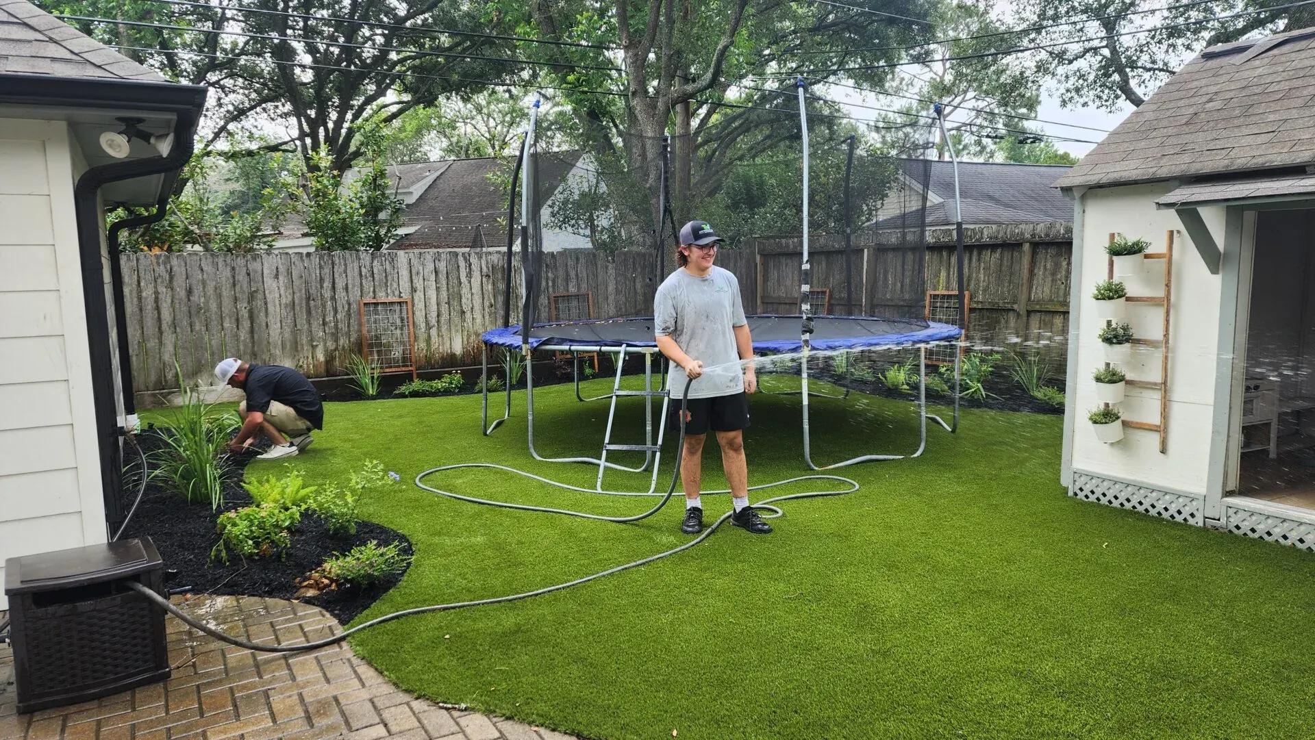 A boy rinsing an artificial turf lawn with a hose beside a trampoline